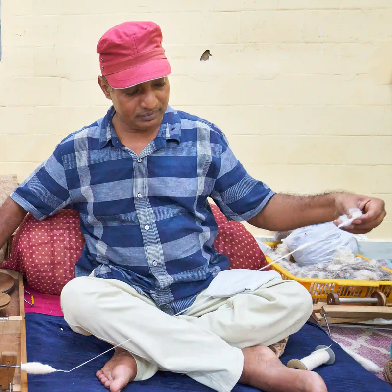 Surjeet, wearing a red cap, sitting cross-legged and spinning wool into yarn