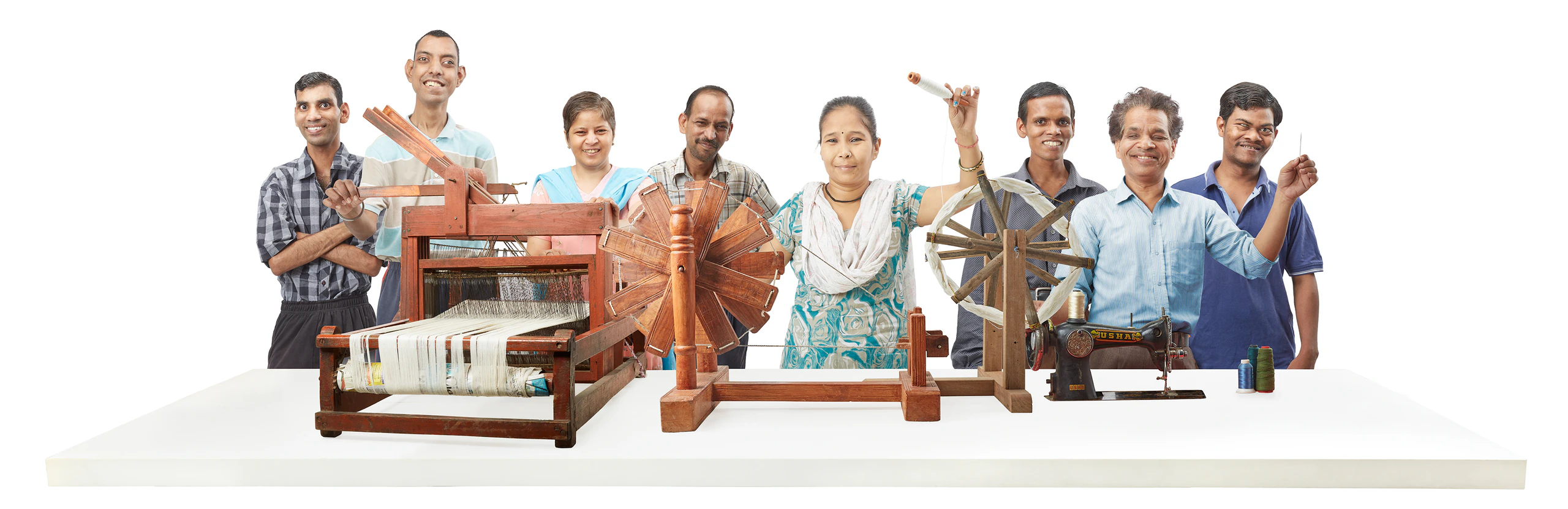 A group of eight smiling artisans standing behind a hand loom, spinning wheels and a sewing machine