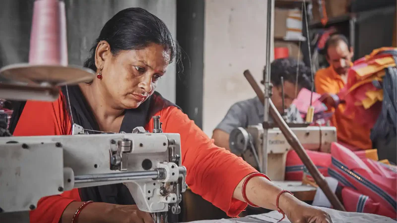 A woman concentrating as she stitches fabric on an industrial sewing machine, with colleagues working behind her